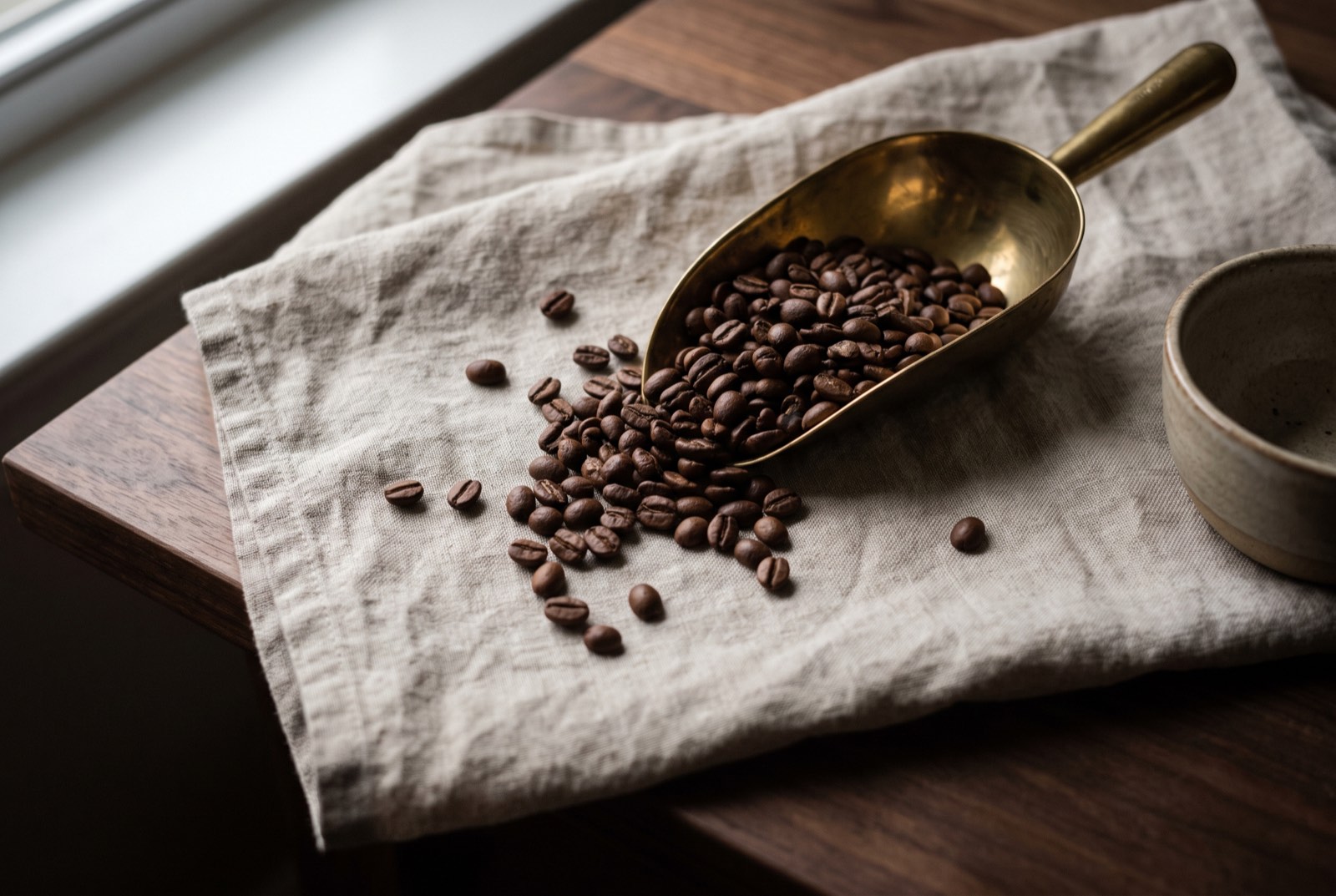 Roasted beans spilling from a brass scoop onto raw linen